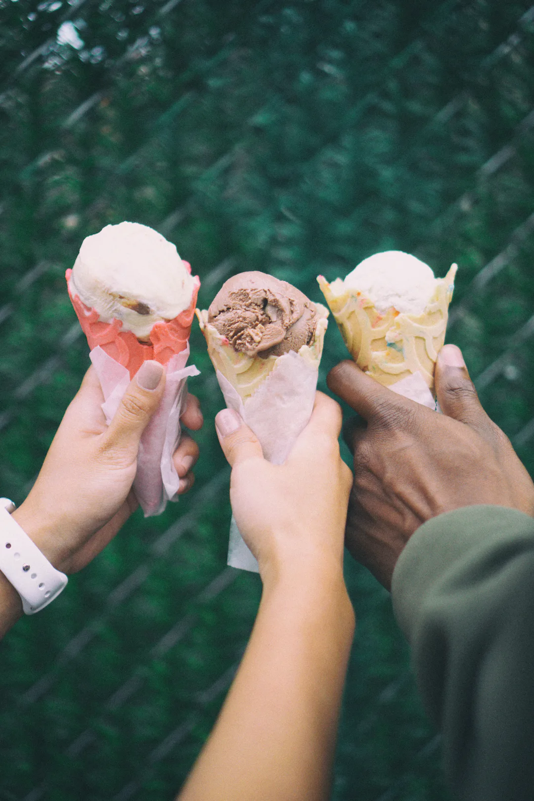 Three hands holding ice cream cones with vanilla, chocolate, and rainbow sprinkle flavors against a blurred green background.