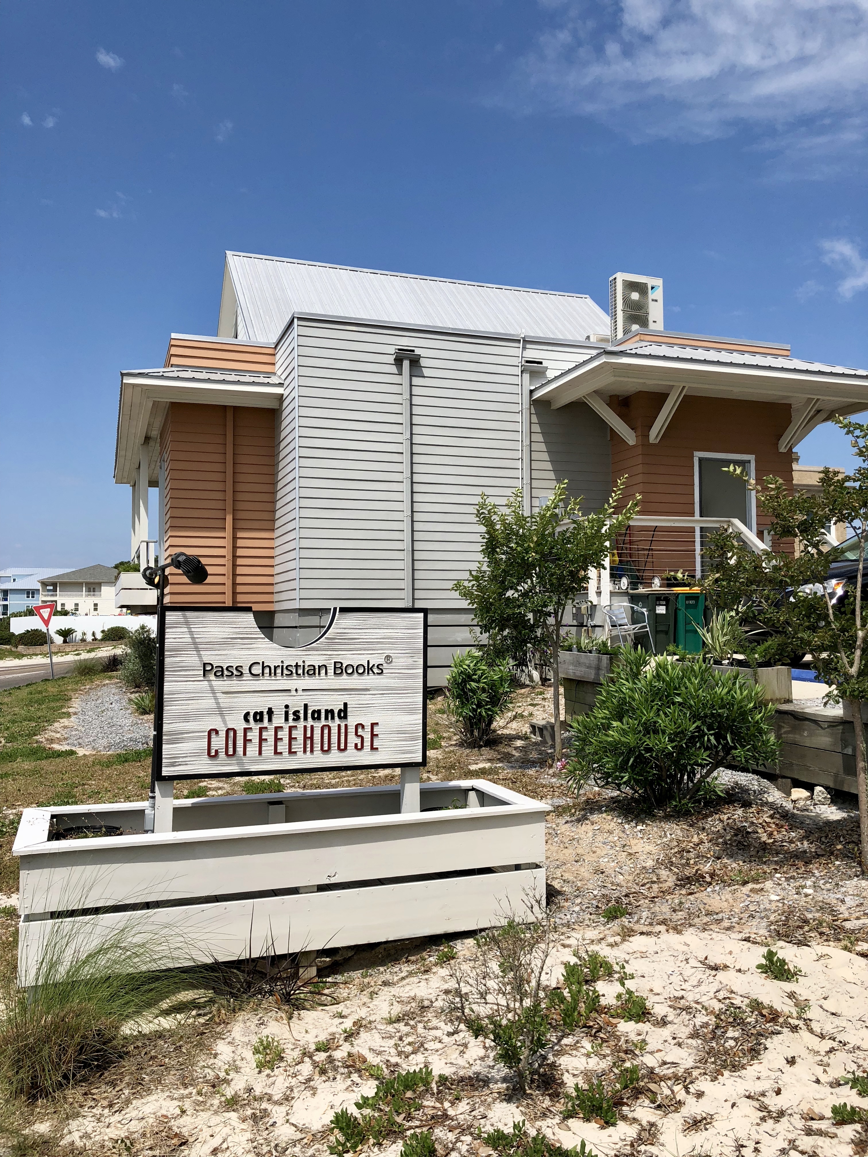 A coastal building with gray and orange siding next to a sign reading 'Pass Christian Books cat island COFFEEHOUSE' under a blue sky.