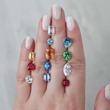 Hand with pastel purple nail polish and small white flower designs, adorned with various colorful gemstones arranged vertically on each finger.
