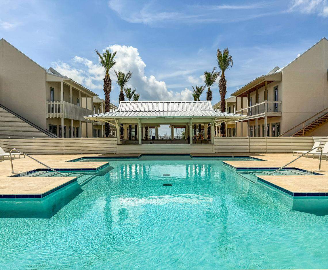 Clear turquoise outdoor swimming pool with lounge chairs, flanked by two beige buildings and palm trees under a blue sky.