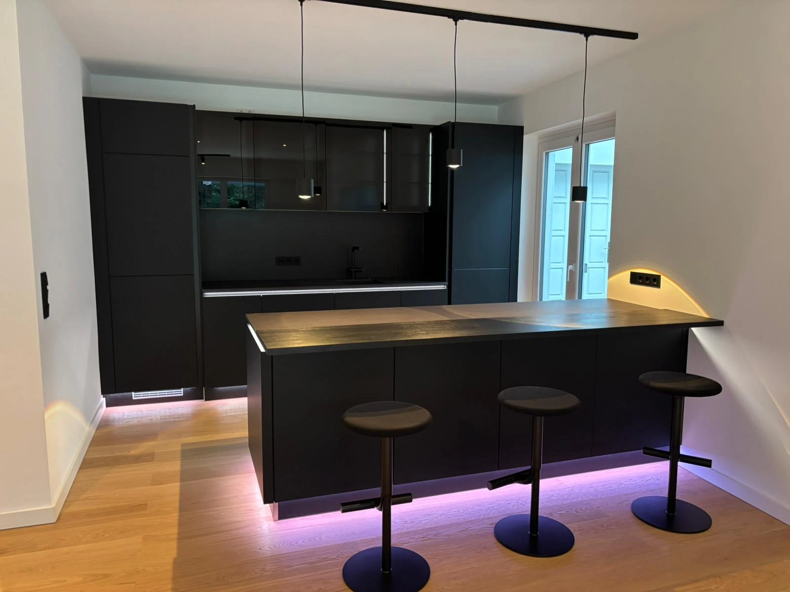 Modern kitchen with black cabinets, a wooden countertop island, three black bar stools, and purple under-cabinet lighting.
