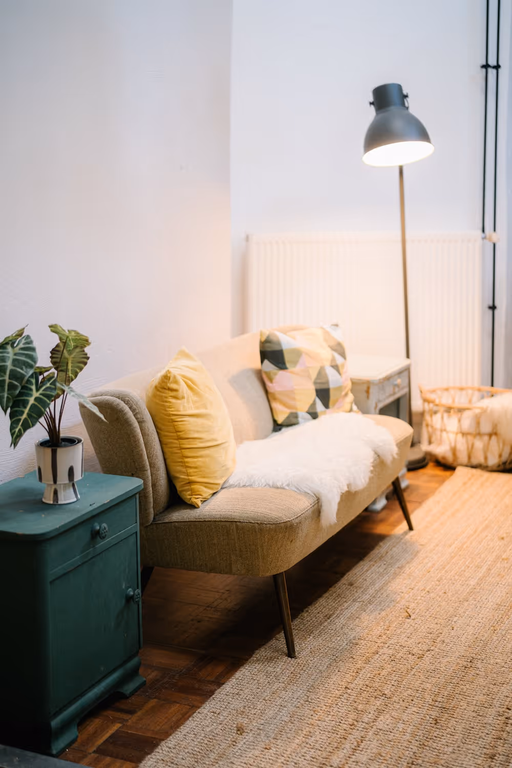 Gemütliches Wohnzimmer mit grauem Sofa, gelben und gemusterten Kissen, Stehlampe und grünem Beistelltisch mit Pflanze.