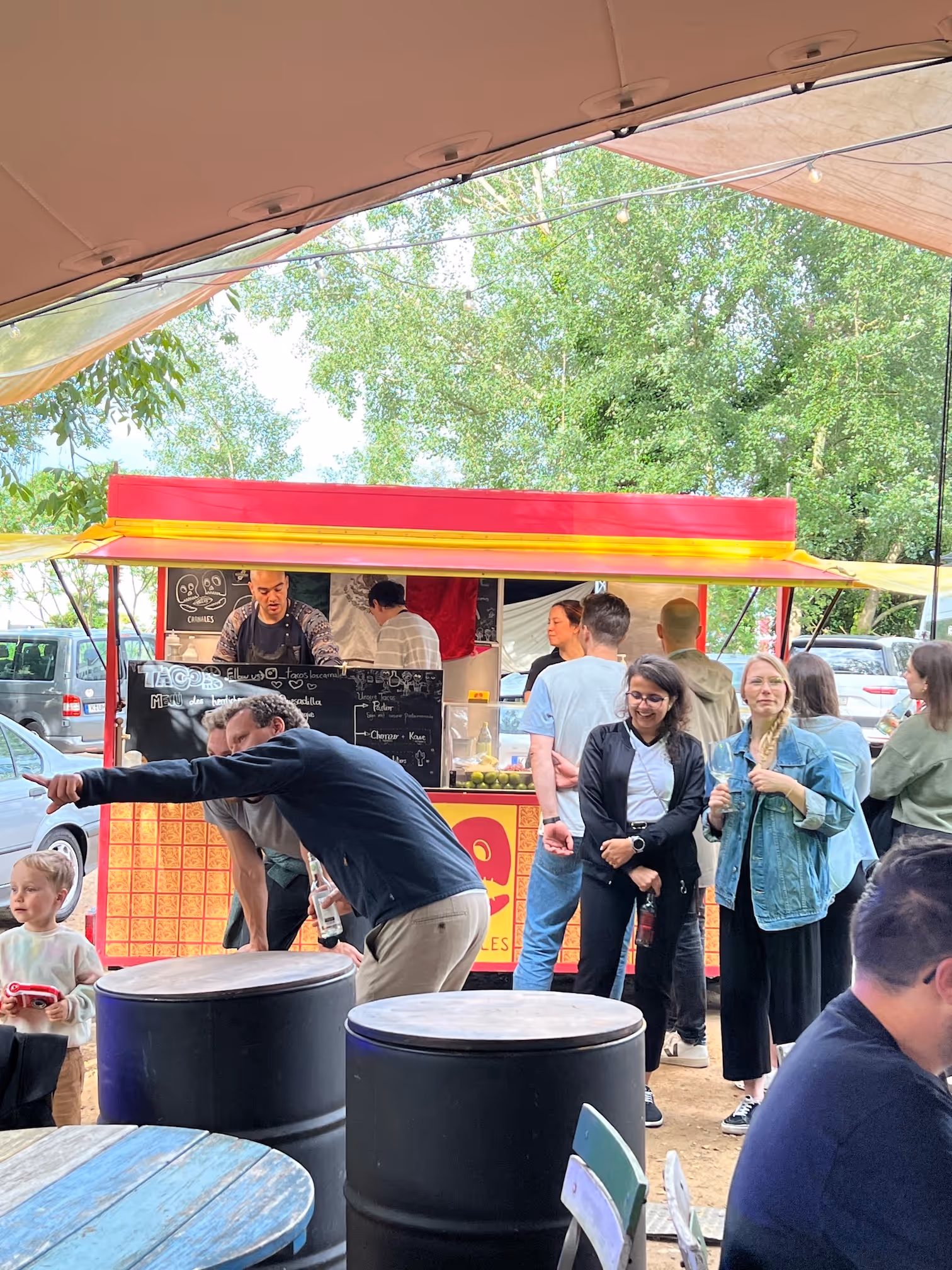 Menschen warten an einem roten und gelben Taco-Stand im Freien unter einem Zelt bei Tageslicht.