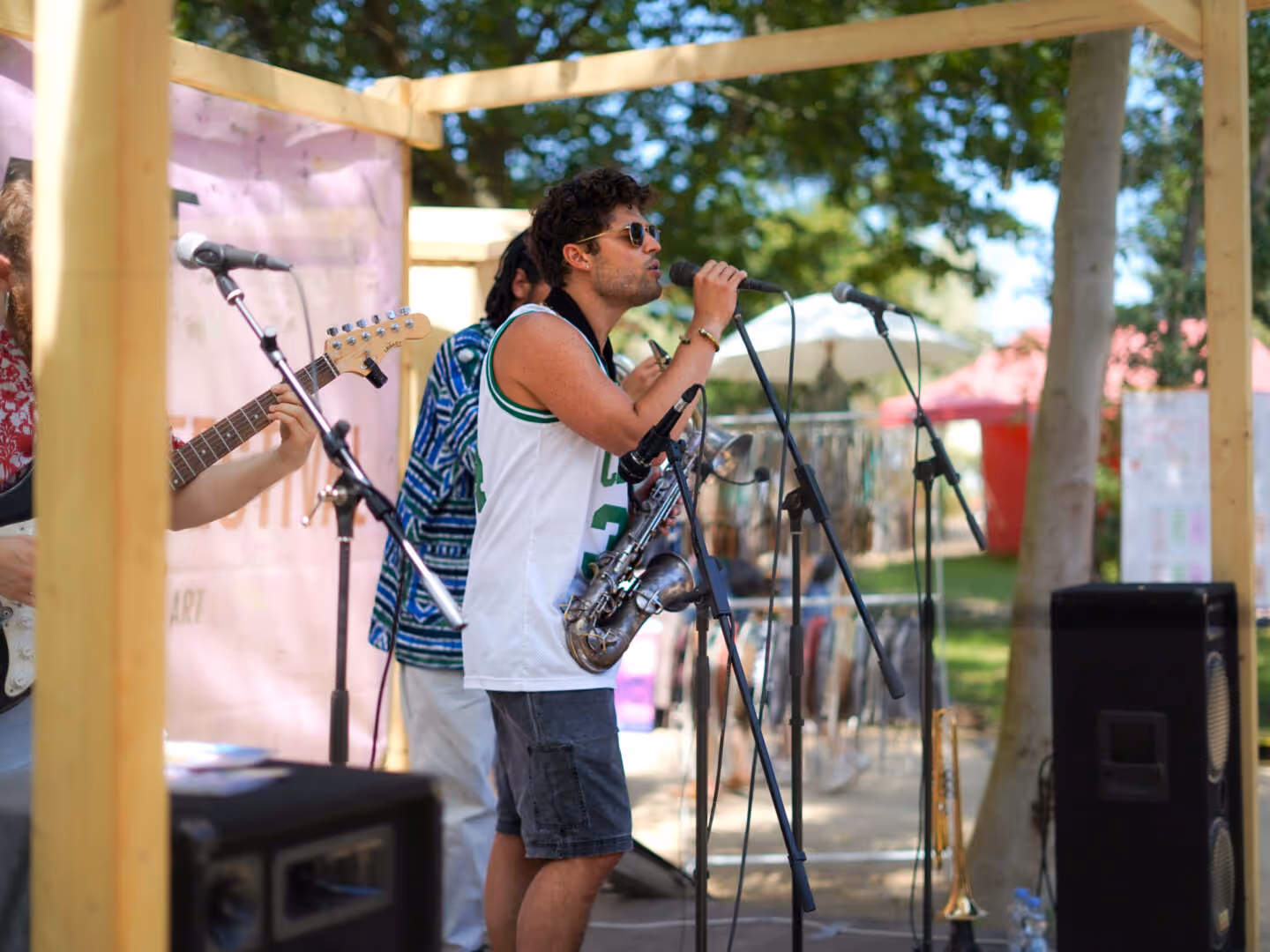 Ein Sänger mit Sonnenbrille und einem Saxophon singt in ein Mikrofon auf einer Bühne im Freien, begleitet von Musikern mit Gitarre und Trompete.