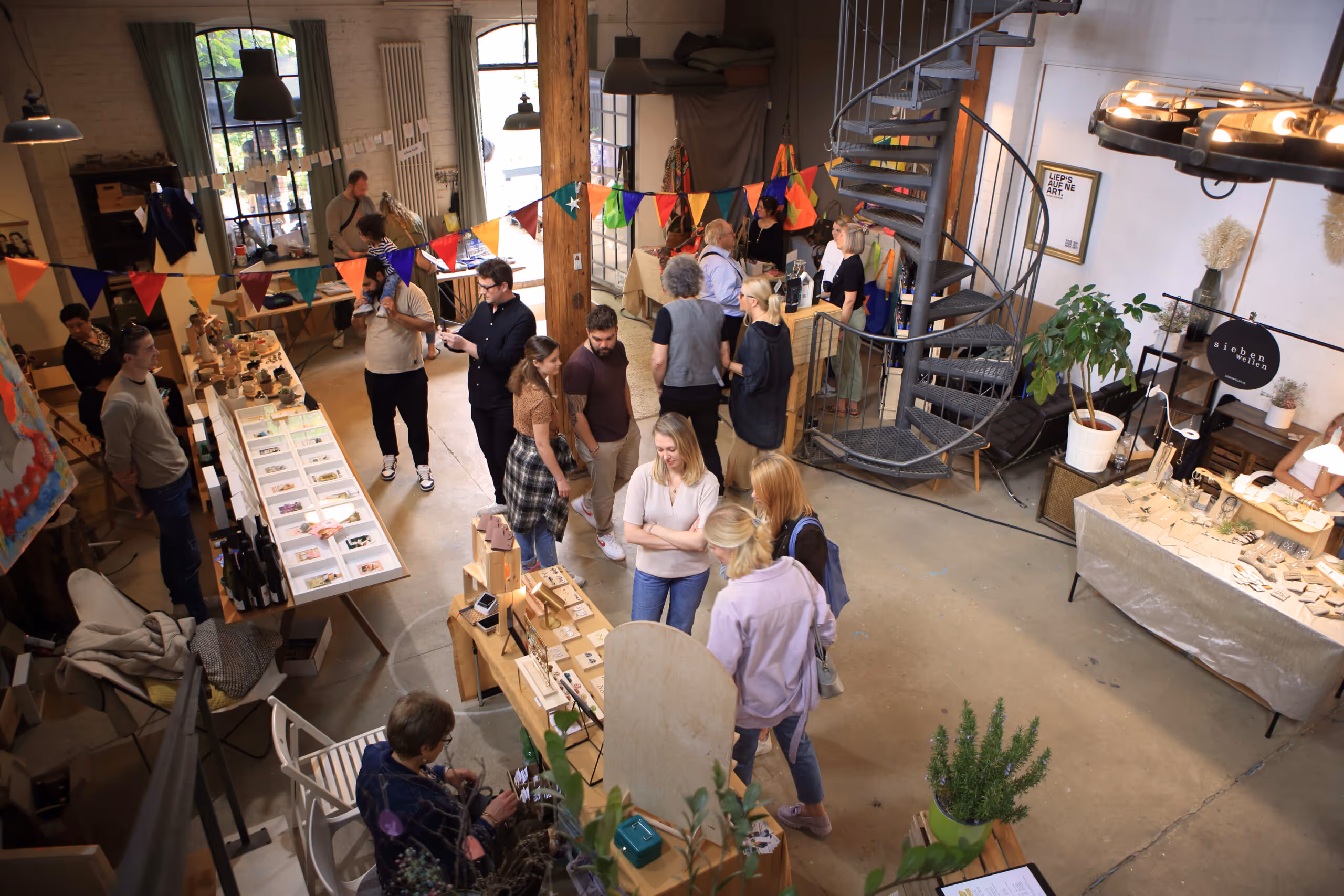 Innenaufnahme eines belebten Handwerks- und Kunstmarkts mit Ständen, die Schmuck, Kunst und Accessoires verkaufen, sowie einer Wendeltreppe in einer Loft-ähnlichen Umgebung.