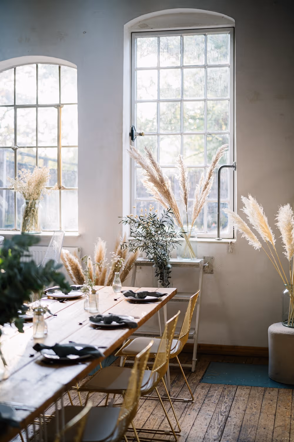 Hell eingerichteter Esstisch mit bunten Stühlen, dekoriert mit Gläsern und getrockneten Gräsern vor großen Fenstern.