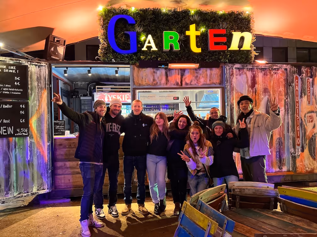 Eine Gruppe von neun fröhlichen Menschen posiert vor einem beleuchteten Schild mit dem bunten Wort ‚Garten‘ an einem Abend.