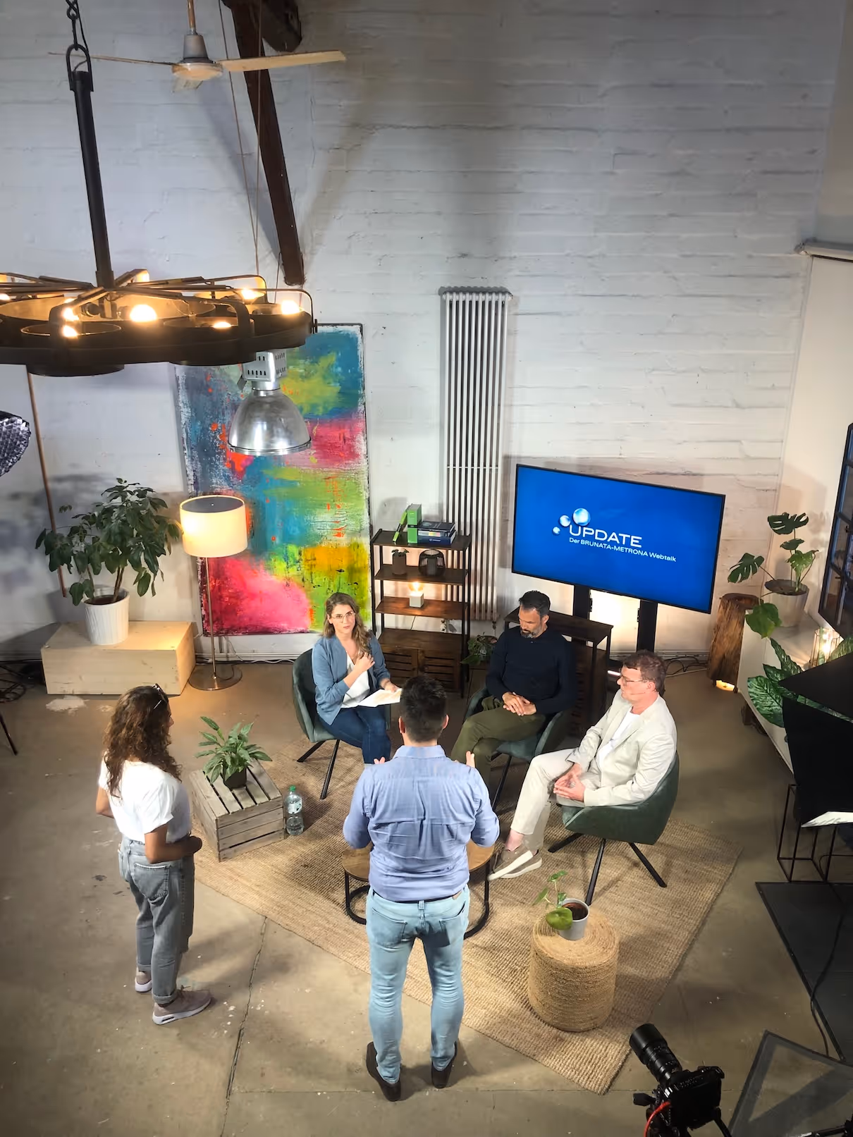 Fünf Personen diskutieren in einem modernen Studio mit hellem Backsteinmauerhintergrund, Pflanzen und einem Bildschirm mit der Aufschrift ‚UPDATE Der BRUNATA-METRONA Webtalk‘.