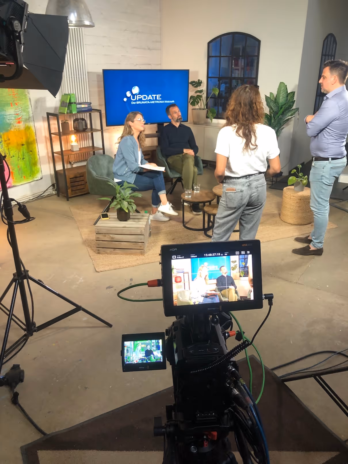 Fernsehstudio mit zwei sitzenden Moderatoren vor einem Bildschirm mit 'UPDATE Der BRUNATA-METRONA Webtalk' und zwei stehenden Personen im Hintergrund.