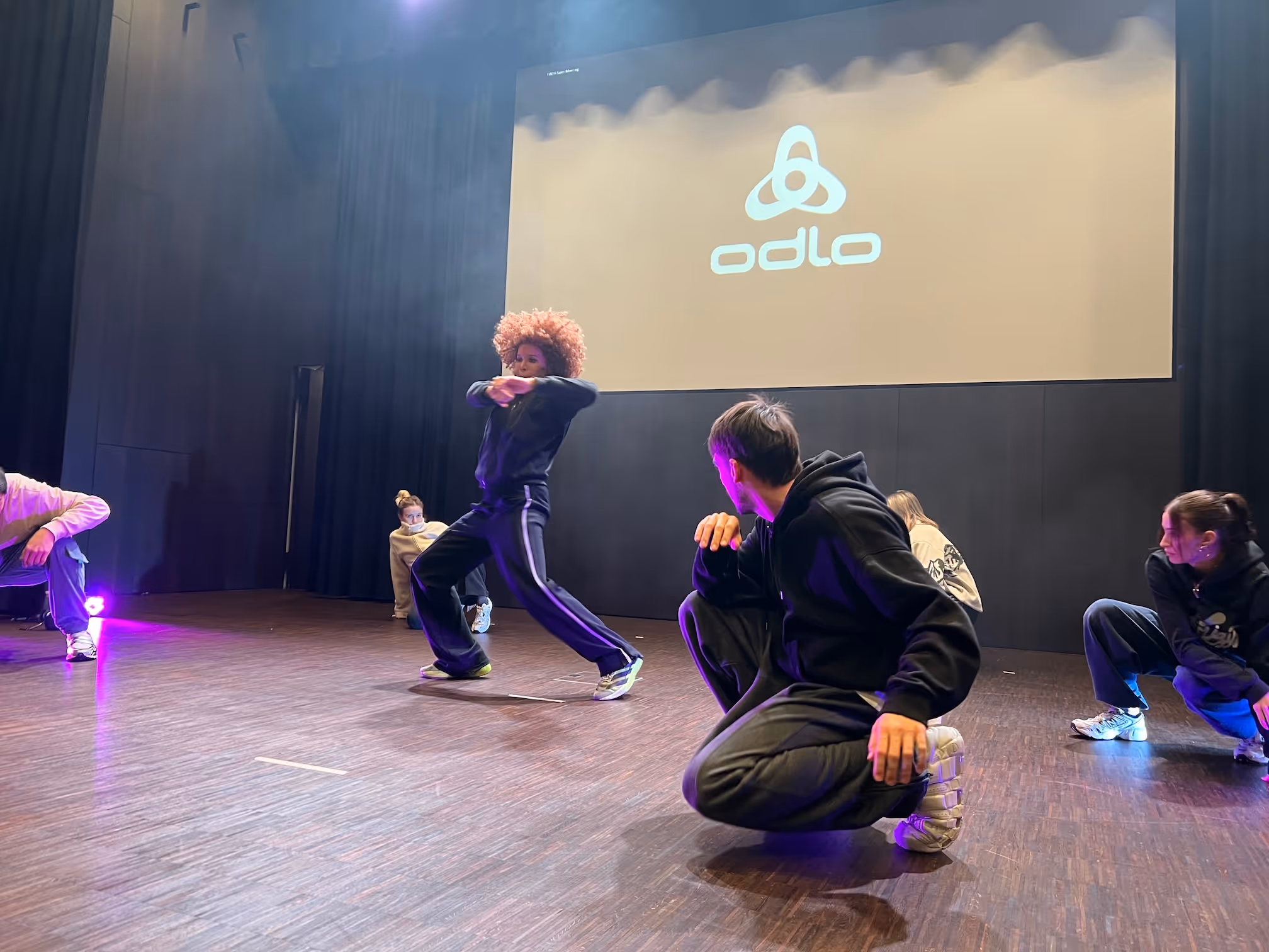 Fünf Tänzer in lässiger Sportkleidung üben Choreografie auf einer Bühne mit einem großen Projektionsbildschirm im Hintergrund.