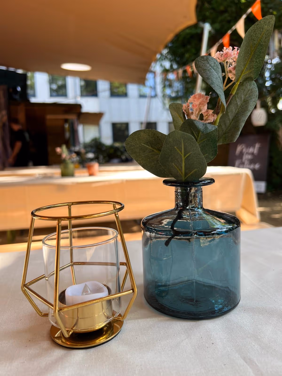 Eine Tischdekoration mit einem blauen Glasvasen mit grünen Blättern und rosa Blüten sowie einem goldfarbenen Kerzenhalter mit einer LED-Teelichtkerze auf einem weißen Tisch.