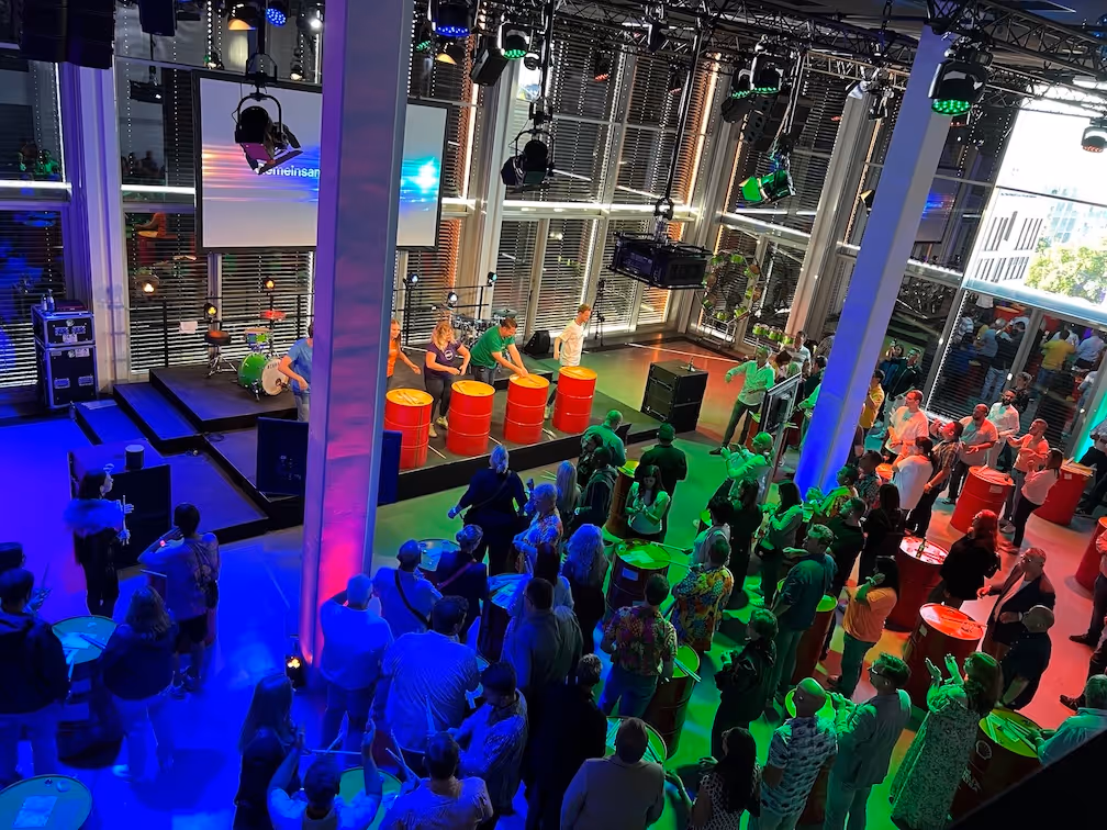 Menschen versammeln sich in einem modernen Veranstaltungsraum mit bunter Beleuchtung, während fünf Personen auf einer Bühne mit roten Fässern eine Performance geben.