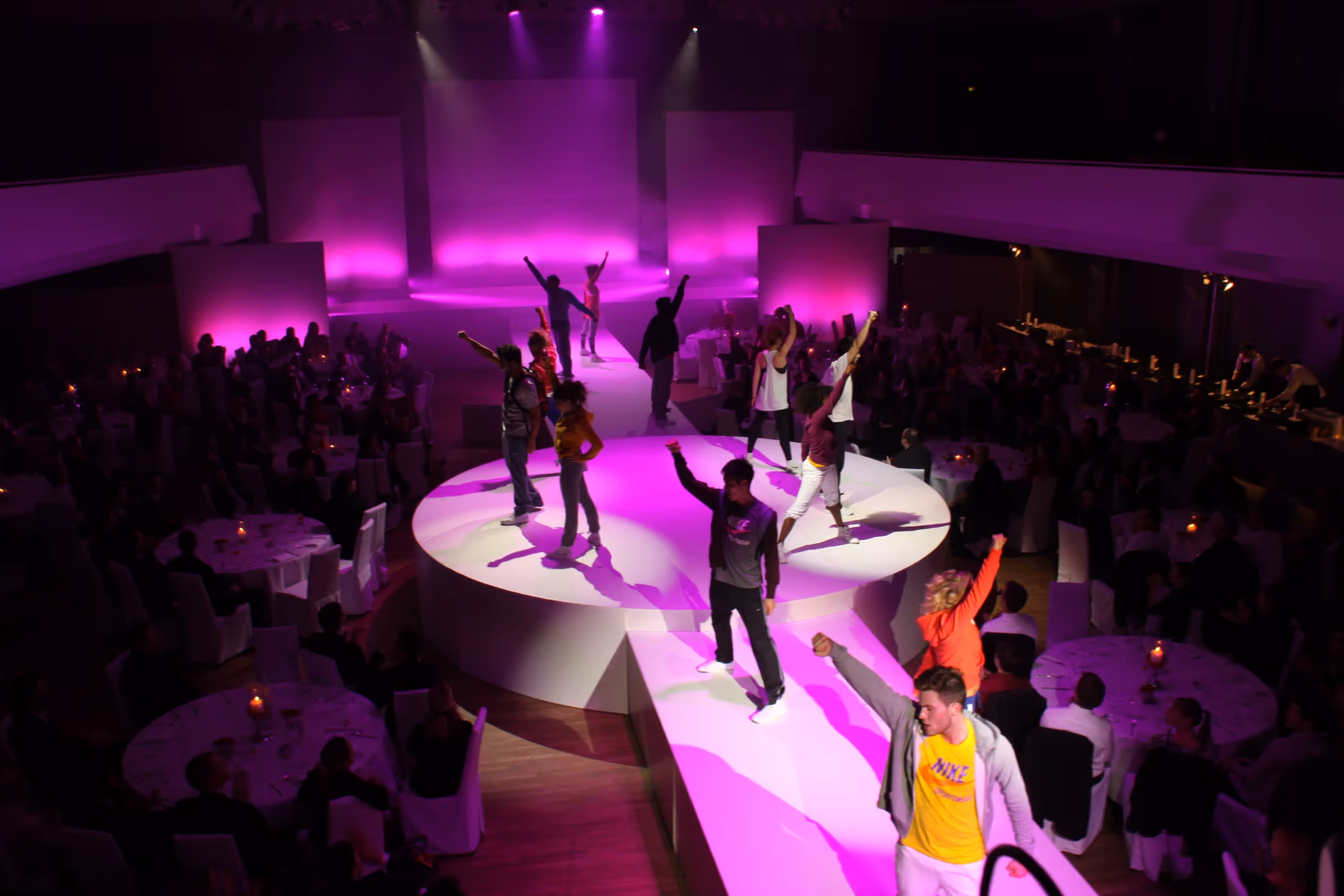 Modenschau mit Models auf einem runden Laufsteg, beleuchtet von lila Bühnenlicht und umgeben von einem Publikum an runden Tischen.