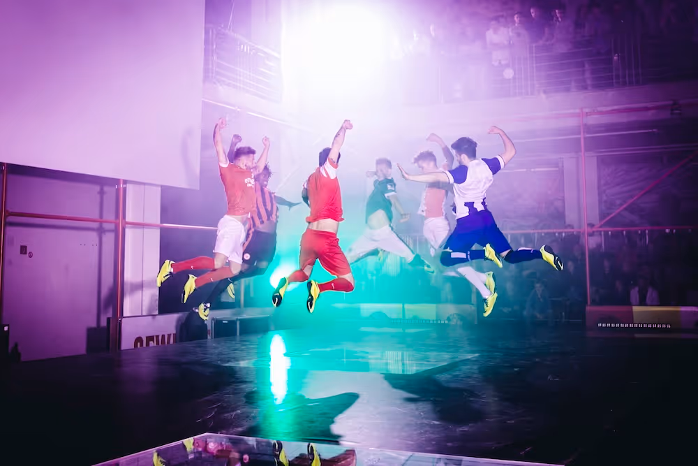 Mehrere Fußballspieler in bunten Trikots springen gleichzeitig in einer Halle mit Zuschauern und buntem Licht.