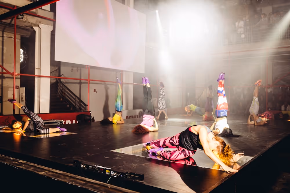 Gruppe von Tänzerinnen auf einer Bühne in sportlicher Kleidung, die verschiedene Stretch- und Balanceübungen ausführen.