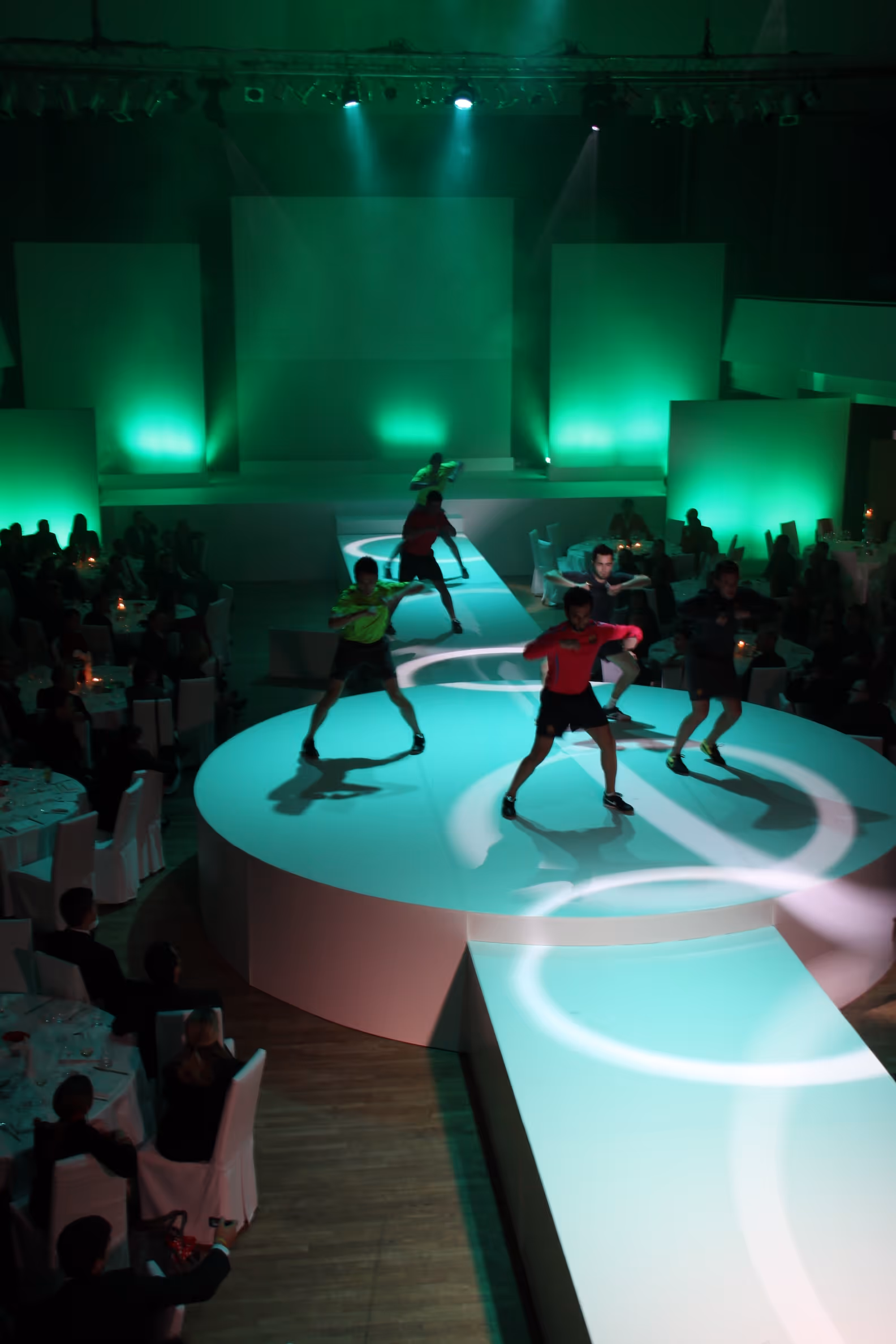 Fünf Männer tanzen auf einer beleuchteten Bühne mit grünem Licht, umgeben von Publikum an eingedeckten Tischen.