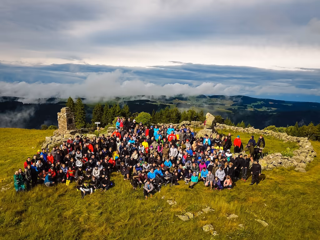 Große Gruppe von Menschen, die auf einer grünen Bergwiese sitzen und stehen, umgeben von alten Steinmauern und Bäumen unter bewölktem Himmel.