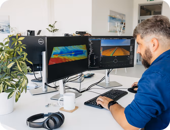 A male engineer analyzing subsea pipeline data and 3D bathymetric seabed models using specialized software at a dual-monitor workstation in the Hidrocibalae office.