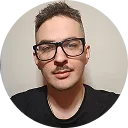 Portrait of a young man with short dark hair, glasses, a mustache, and a slight beard wearing a black shirt against a plain background.