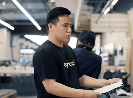 Man in a black eytap t-shirt holding and looking at a document inside an industrial or workshop setting.