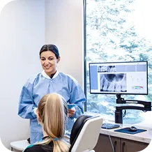 Female dentist in blue scrubs smiling and talking to a seated patient with dental X-rays displayed on a monitor.