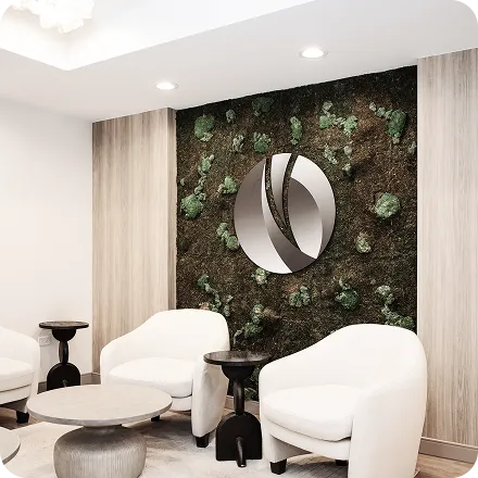 Modern waiting area with white armchairs, small black side tables, a round coffee table, and a green moss wall featuring a large circular metallic logo.