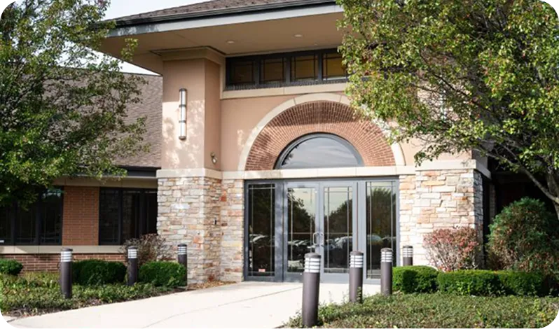 Building entrance with double glass doors, stone and brick facade, surrounded by trees and landscaping.