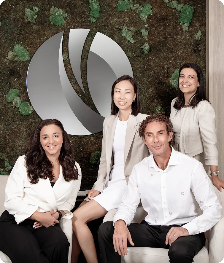 Group of four smiling professionals seated and standing indoors in front of a green moss wall with a large silver abstract logo.