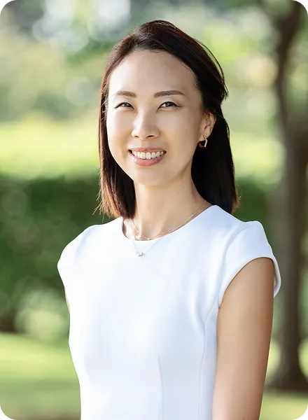 Smiling woman with straight dark hair wearing a white sleeveless top standing outdoors with blurred greenery in the background.