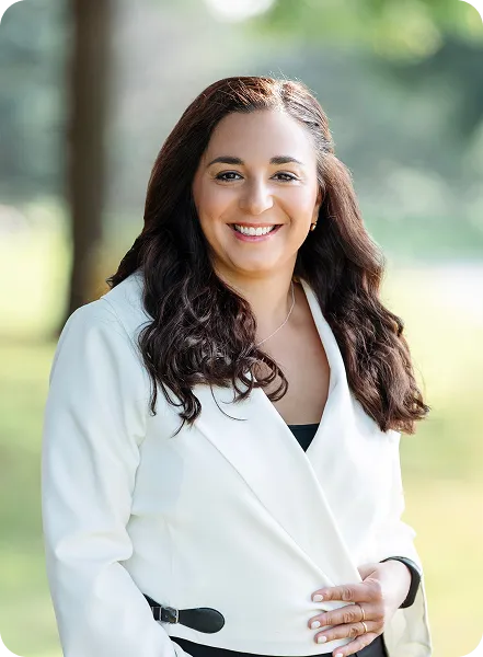 Smiling woman with long dark hair wearing a white blazer standing outdoors with blurred trees in the background.