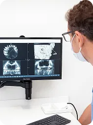 Medical professional wearing glasses and a mask examining 3D dental and jaw X-ray images on a computer screen.