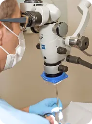 Healthcare professional wearing mask and gloves using a surgical microscope for a precise medical procedure.