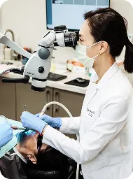 Dentist wearing a mask and gloves examining a patient's teeth using a microscope in a dental clinic.
