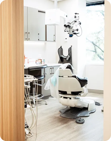 Modern dental office with a white dental chair, overhead dental microscope, and cabinets under natural light from a window.