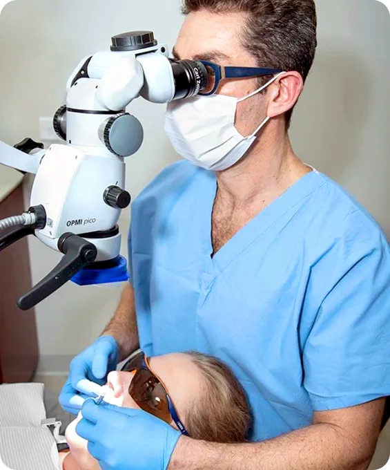 Dentist wearing blue scrubs, mask, and gloves examining a patient’s teeth using a dental microscope.
