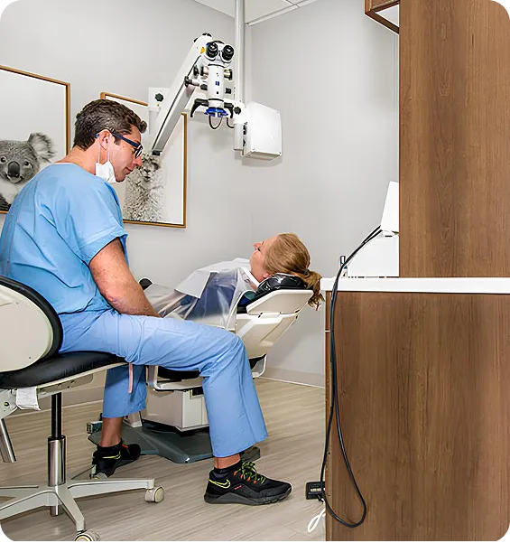 Dentist in blue scrubs and mask seated next to a patient reclining in a dental chair in a modern clinic room.