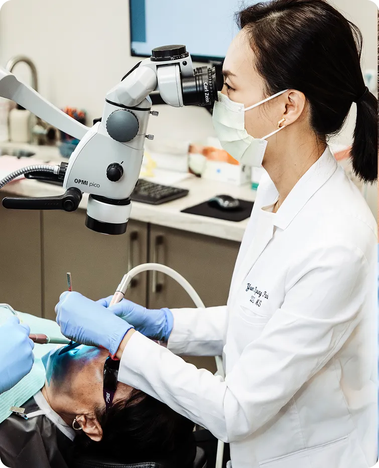 Dentist wearing a mask and gloves using a dental microscope while treating a patient lying in a dental chair.