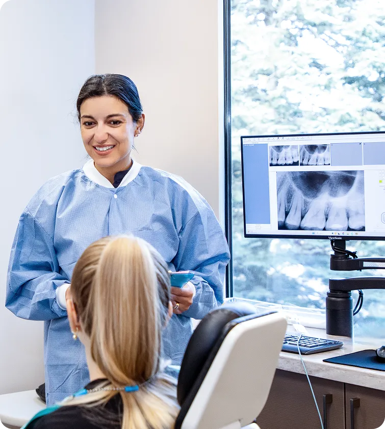 Dentist in blue protective gown talking to a seated female patient with dental X-rays displayed on a monitor beside them.