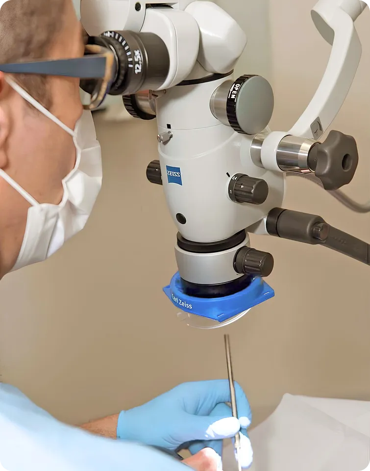 Medical professional wearing a mask and blue gloves using a Zeiss microscope during a procedure.