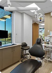 Modern dental treatment room with dental chair, overhead light, computer, and window showing outdoor view.