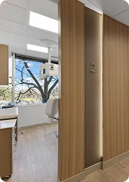 Modern dental clinic room with wooden sliding door labeled number 2, dental chair, window showing bare tree outside.