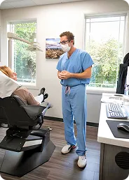 Dentist in blue scrubs and face mask talking to patient reclining in dental chair by two large windows.