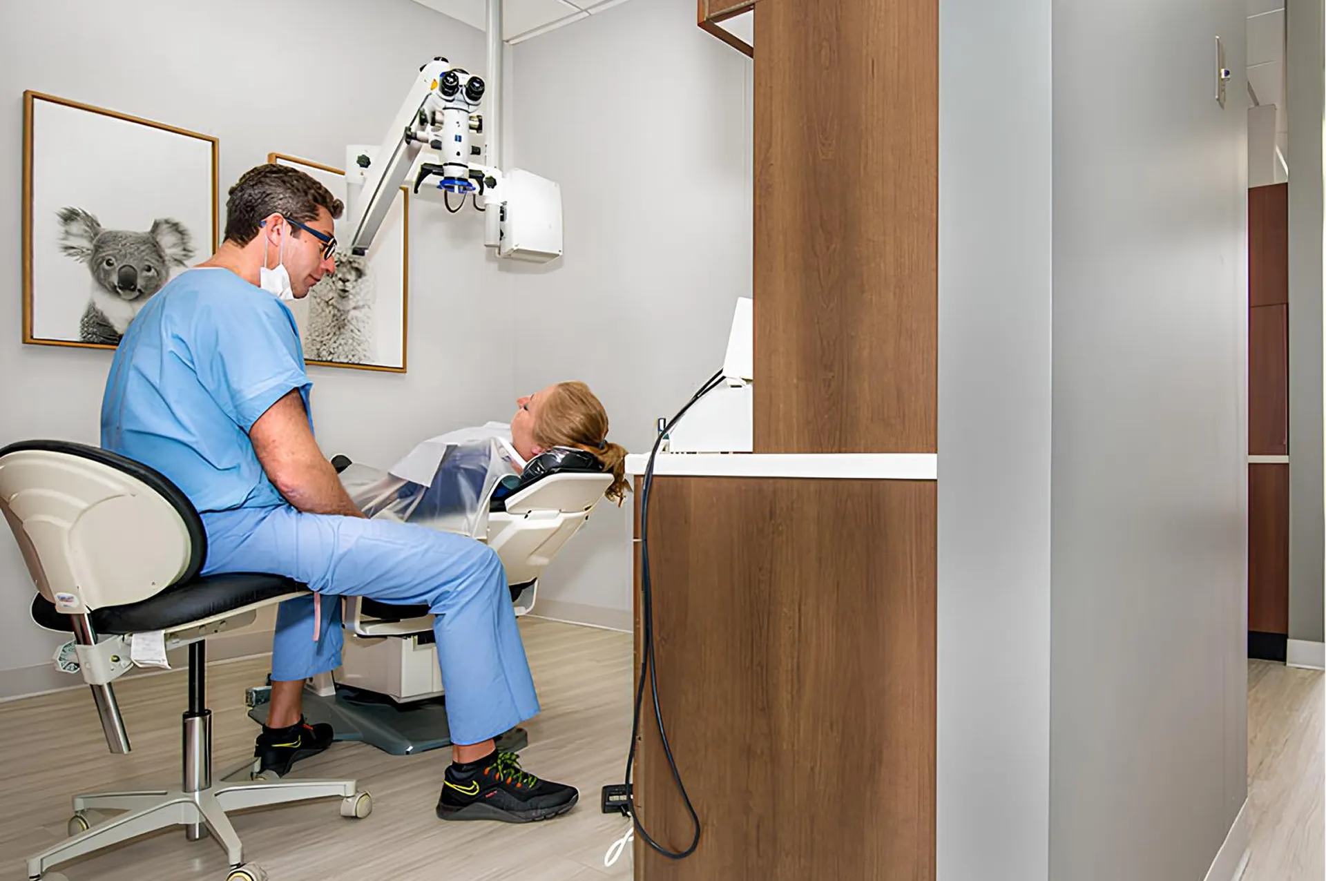 Dentist in blue scrubs sitting on a chair next to a reclining patient in a dental office with microscope equipment and koala pictures on the wall.