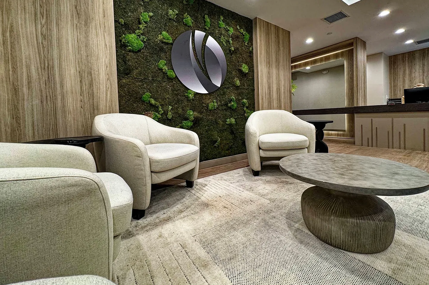 Modern lounge area with three beige armchairs, a round stone coffee table, moss-covered wall with a circular logo, and a wooden-paneled reception desk.