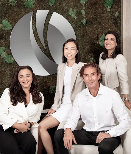 Group portrait of two men and two women in business attire seated and standing in front of a green moss wall with a large abstract silver logo.