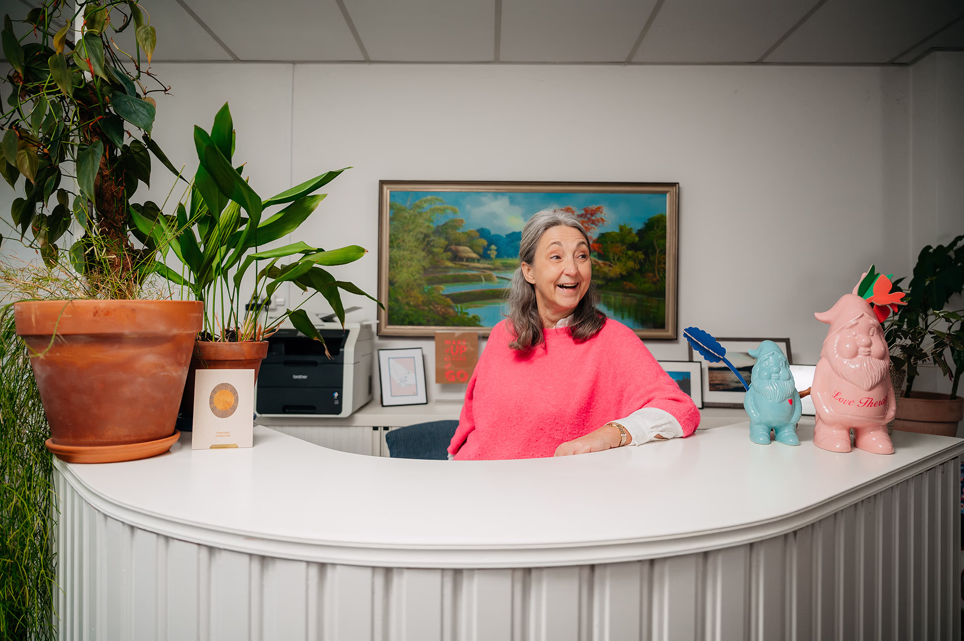 Vrouw met roze trui kijkt lachend opzij achter een witte houten balie