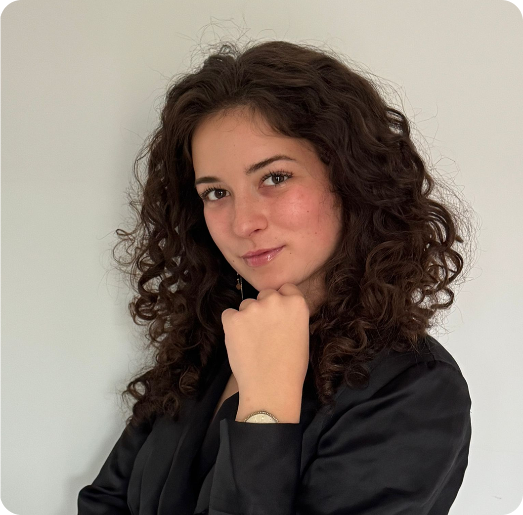 Young woman with curly dark hair wearing a black blazer, posing with her hand under her chin against a plain light background.