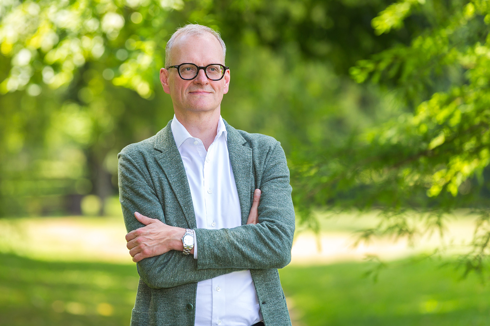 Middle-aged man wearing glasses and a gray blazer standing outdoors with arms crossed, surrounded by greenery.