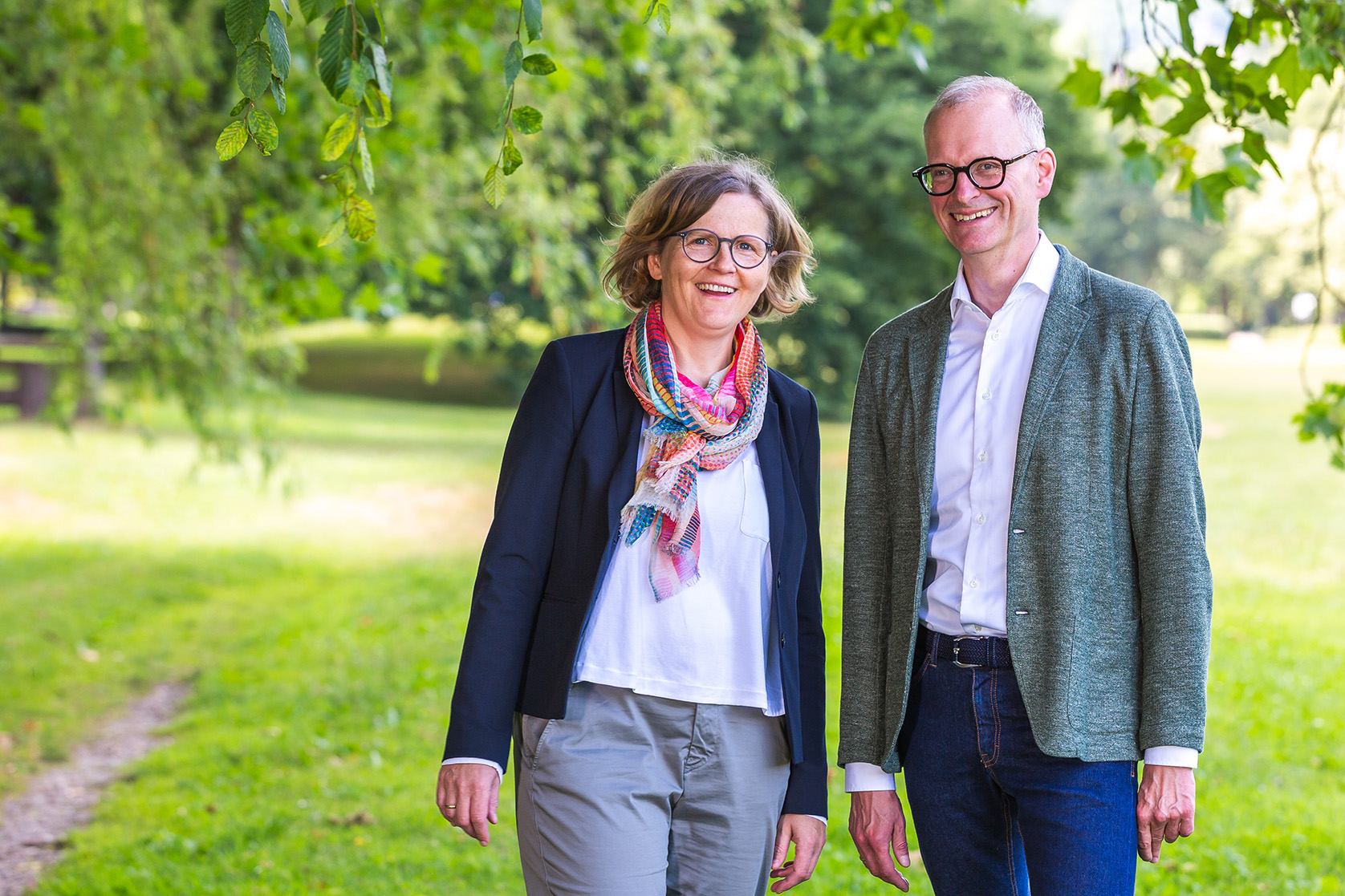 Smiling man and woman standing outdoors with green trees and grass in the background.