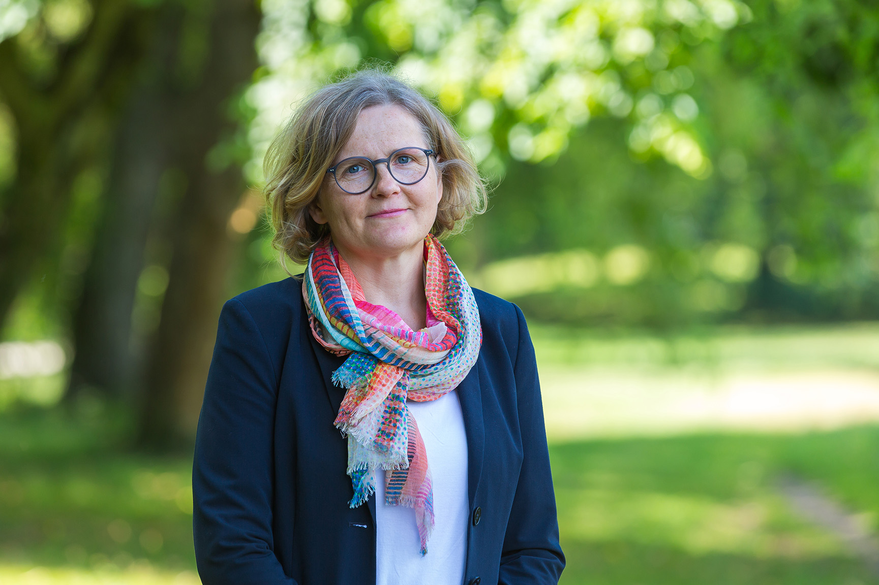 Woman with glasses wearing a colorful scarf and dark jacket standing outdoors with green foliage background.